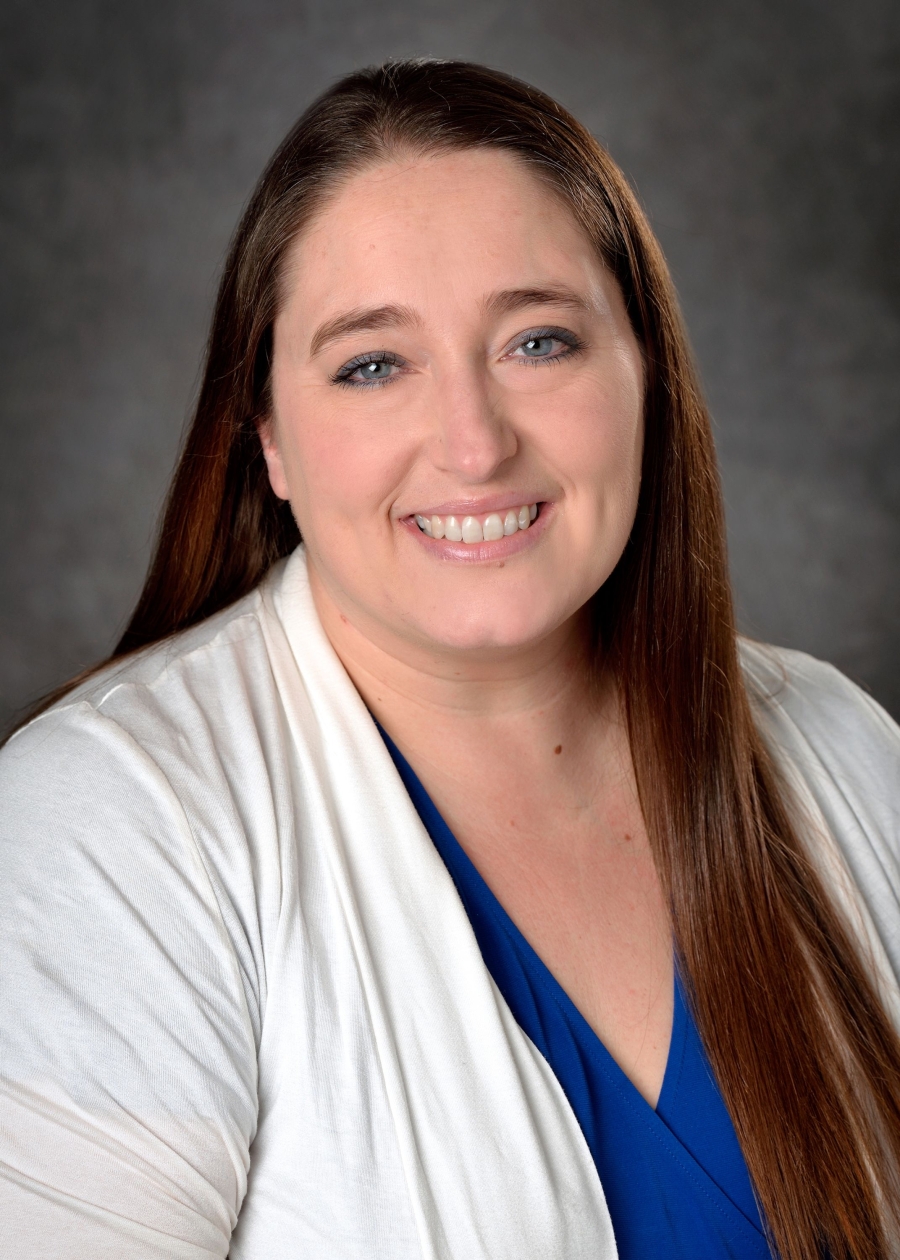 Headshot of Shelby Gombosi, Caucasian woman with long, brown hair wearing a dark blue blouse and a white cardigan sweater.