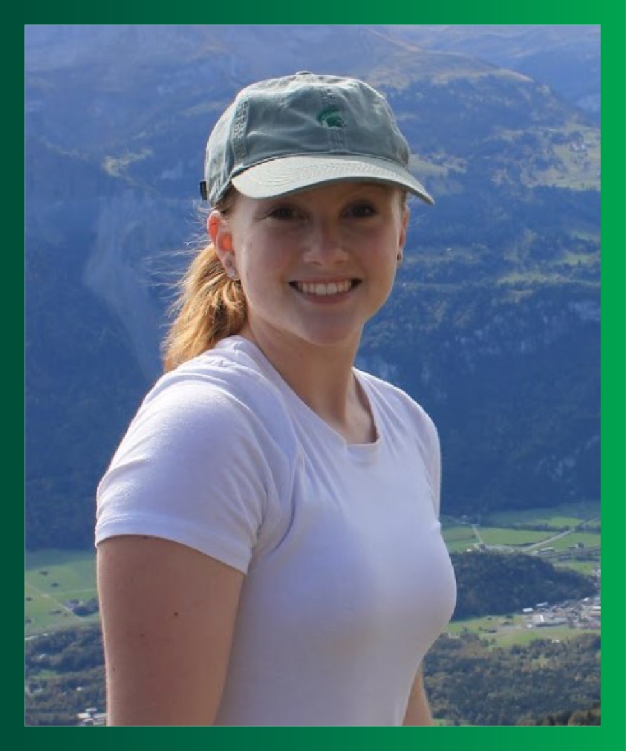 Picture of Kellie W., smiling Caucasian female, wearing a white t-shirt, green baseball cap, and standing in front of mountains.