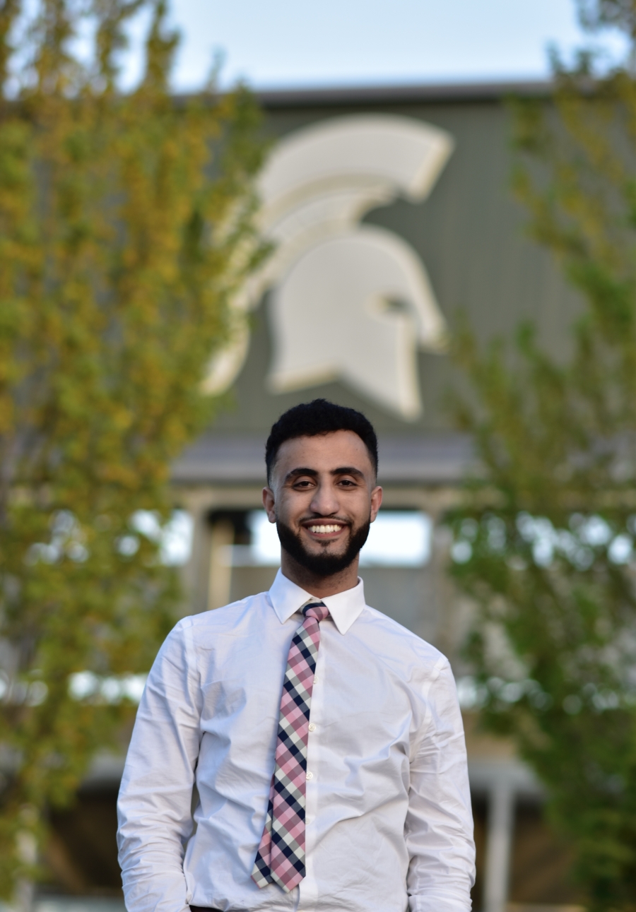 Photo of Kareem standing outside with the Spartan head logo on the big screen of the football stadium in the background. Kareem is wearing a white button-down shirt with tie, has short black hair and full beard, who is smiling.