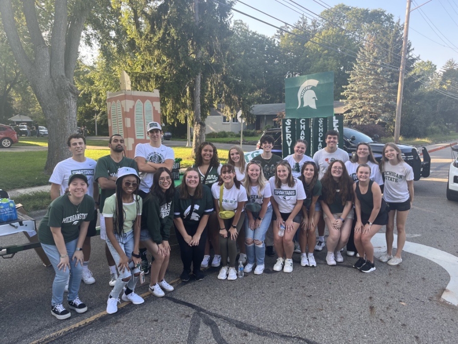 Group of 20 students posing in the middle of a road in front of a parade float featuring the Beaumont Tower made of cardboard smiling at the camera on a sunny day.