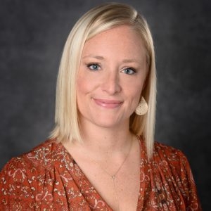 Photo of Tiffany smiling and wearing a v-neck, reddish orange blouse with necklace. Tiffany has shoulder length blonde hair, parted to one side.