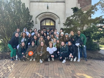 Group of 50 students in front of Beaumont Tower in workout clothes and jackets smiling at the camera on a cloudy day