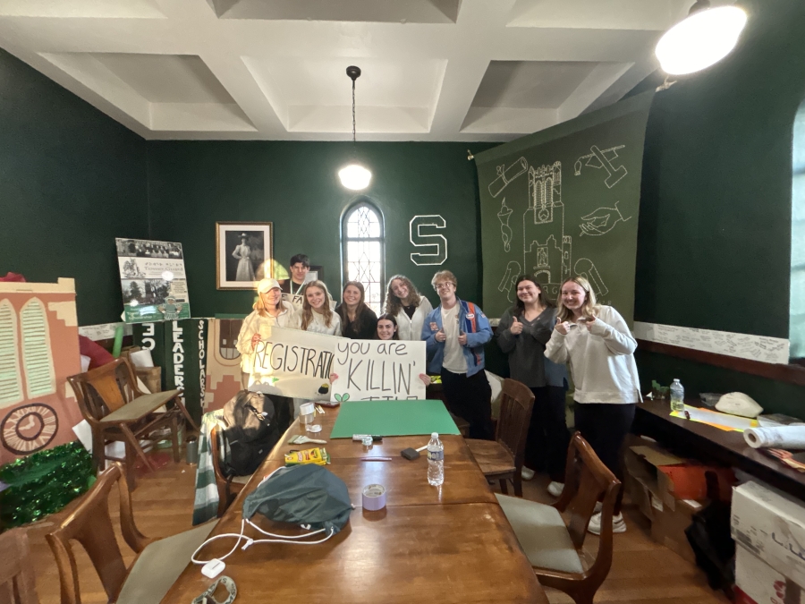 Group of 9 students standing inside a green room within Beaumont Tower behind a table holding encouraging signs, some giving a thumbs up, and smiling at the camera