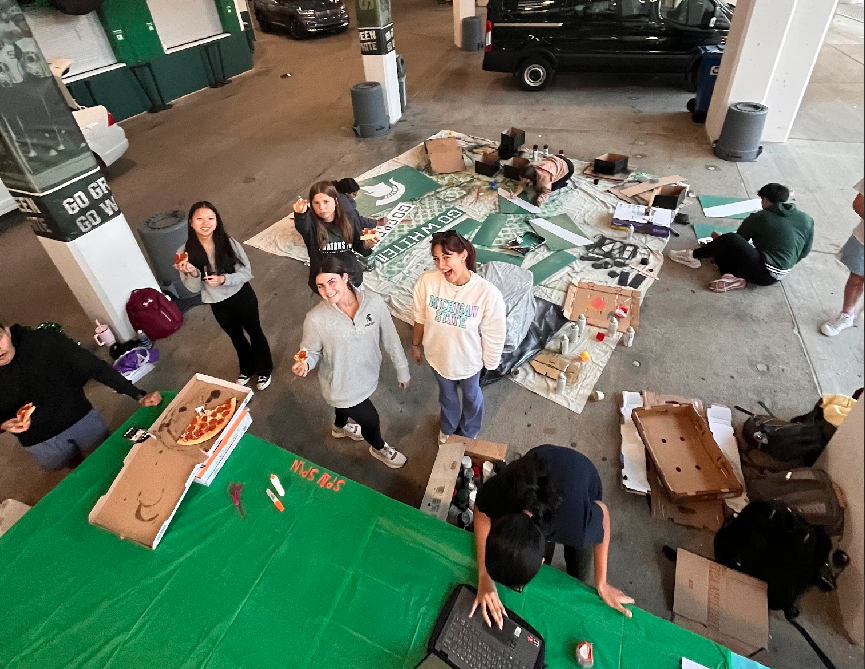 Picture taken from a higher perspective looking down on students painting Homecoming float signs, typing on a computer, eating pizza, and smiling at the camera