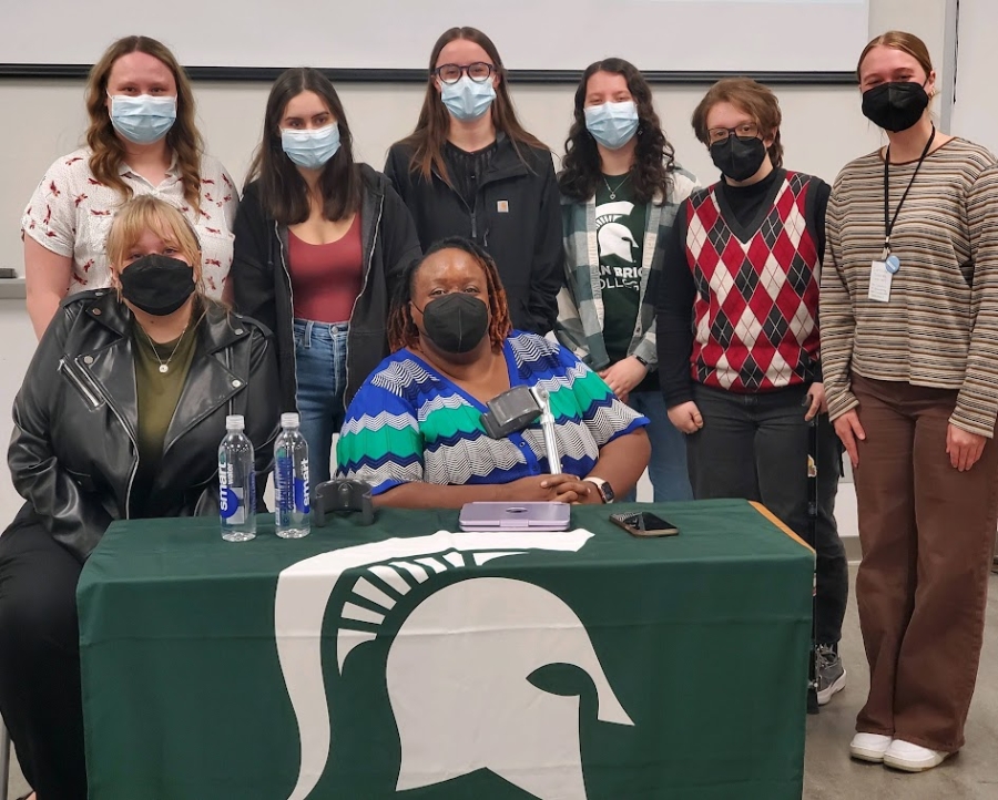 Group of 8 people wearing masks sitting and standing behind a table with a Sparty head symbol on a green table cloth looking towards the camera