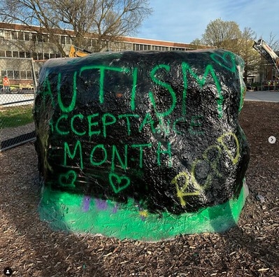 back side of MSU rock spray painted black with writing in green "Autism Acceptance Month" with green hearts and "RCPD" written in gold on the lower right corner and "Tower Guard" written in gold on the lower left corner surrounded by orange hand prints of those who painted the rock