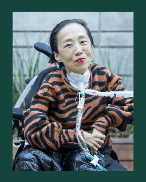 lice is an Asian American woman in a power chair. She is smiling at the camera, wearing an oxygen mask and a tiger striped sweater. 