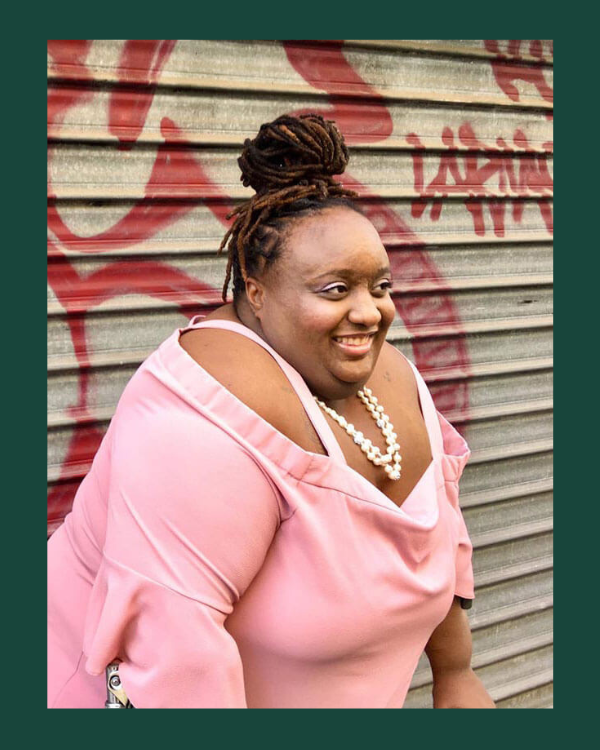 Imani Barbarin is a Black disabled woman smiling in a pink shirt against a garage door with red graffiti.