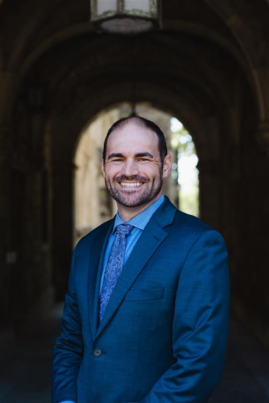 Allen is smiling and wearing a blue suit and coordinating tie. He has short black hair and facial hair.