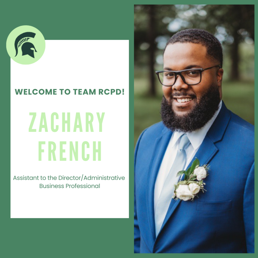 Zach is smiling, has short black hair and facial hair and is wearing a blue suit and tie. He is standing outside. 