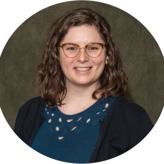 Photo of Tesia Freer- A smiling light skinned woman with shoulder length light brown curly hair and glasses. She is wearing a black cardigan over a blue shirt.