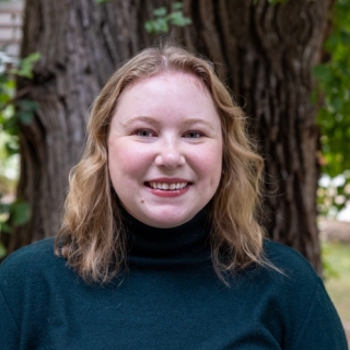 A smiling white woman with shoulder length, blonde hair parted in the middle. She is wearing a green turtleneck sweater.  She is standing in front of a tree with green leaves.  