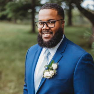 Zach is standing outside, smiling and is wearing a blue suit and tie. He is wearing glasses and has short black hair and facial hair.